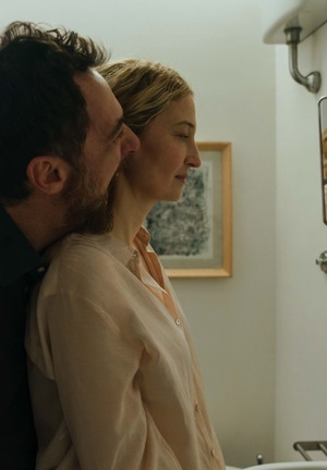 Intymny kadr filmowy z dramatu &amp;amp;amp;quot;Trzy pożegnania&amp;amp;amp;quot;. Mężczyzna czule przytula kobietę od tyłu w jasnej łazience. Parę widać w lustrze nad umywalką.