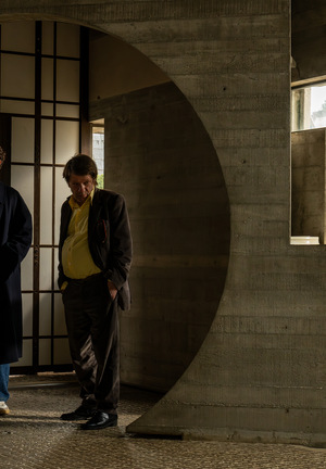 [Kolorowy kadr z filmu. Trzej mężczyźni stoją w okrągłych drzwiach nowoczesnym budynku.]