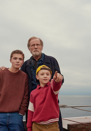 Kolorowe zdjęcie. Dziadek i dwóch wnuków: nastolatek i młodszy chłopiec. Stoją razem na płynącej łodzi, młodszy chłopiec wskazuje na coś palcem, dziadek i nastolatek patrzą w tym kierunku.