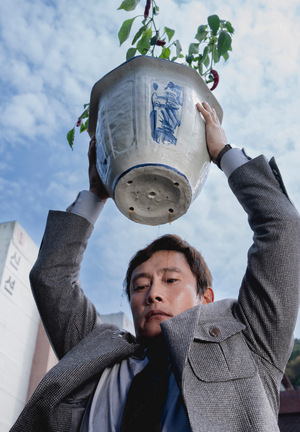 Kolorowy plakat. Mężczyzna w marynarce unosi nad głową doniczkę, którą zaraz ciśnie o ziemię. W tle niebo.