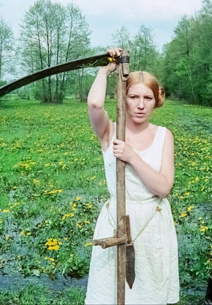 Kolorowy kadr. Kobieta z sierpem na łące. Obok niej mężczyzna w mundurze, patrzy na nią zaintrygowany.