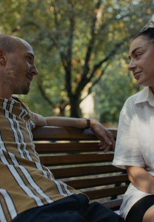 Kolorowe zdjęcie. Dwie osoby, młoda kobieta i młody mężczyzna siedzą na ławce w parku.