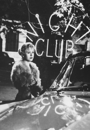 [Czarno-biały kadr z filmu. Zamyślona kobieta stoi przy samochodzie. Za nią widać neon z napisem Night Club.]