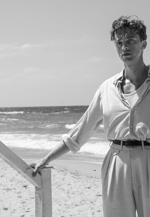 Czarno-biały kadr z filmu. Młody mężczyzna stoi na schodach. Za nim plaża i morze.