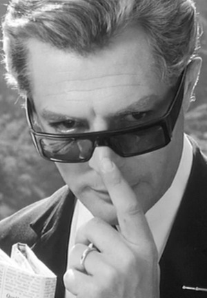 [Czarno-biały kadr z filmu. Mężczyzna zsuwa palcem okulary przeciwsłoneczne i spogląda ponad nimi.]
