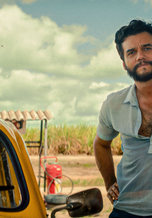 Kolorowe zdjęcie. Mężczyzna z Brazylii opiera się o słup przy drodze. W tle samochód