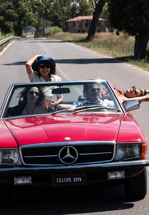 Kolorowy kadr z filmu. Cztery osoby, trzy kobiety i mężczyzna, jadą czerwonym kabrioletem pustą ulicą.