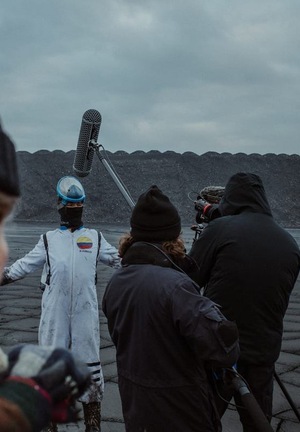 Kolorowe zdjęcie. Lewa część kadru to twarz dojrzałego mężczyzny patrzącego w kamerę. W tle nagrania filmu na pustynnym tle. Osoby w strojach kosmonautów, człowiek z mikrofonem na boomie.