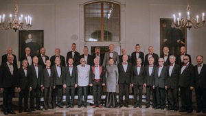 Chór Męski Copernicus - jego członkowie stojący w czarnych garniturach. Stoją w dwóch szeregach w zabytkowej sali jakiegoś pałacu. W tle duże portrety właścicieli pałacu.