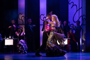 Para w eleganckich strojach tańczy na scenie, w tle siedzą i stoją inni uczestnicy sceny w teatralnej aranżacji.