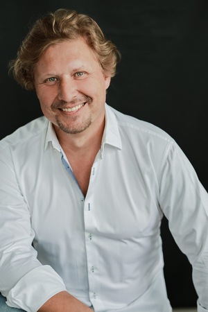 Uśmiechnięty mężczyzna w jasnej koszuli z rozpiętym i górnymi guzikami. Ma jasne włosy i zarost.