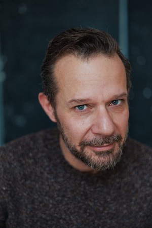 Portret uśmiechniętego mężczyzny w szarym swetrze na zielonym tle.