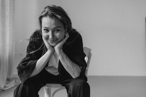 Czarno-biała fotografia uśmiechniętej, młodej kobiety. Siedzi na krześle, rękami opartymi na kolanach podtrzymuje głowę. Ma długie, rozpuszczone włosy.