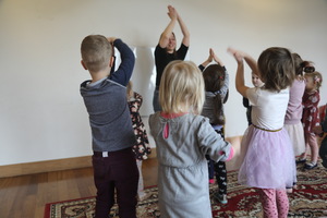 Grupa małych dzieci stoi w sali i wykonuje ruchy z uniesionymi rękami, naśladując prowadzącą stojącą przed nimi. Zajęcia odbywają się w jasnym pomieszczeniu z drewnianą podłogą i dywanem.