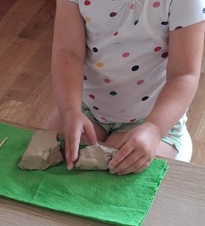 Dziewczynka siedzi przy stole i lepi z gliny na zielonej podkładce. W dłoniach trzyma kawałek jasnej masy, obok leżą inne fragmenty. Ubrana w koszulkę w kolorowe kropki.
