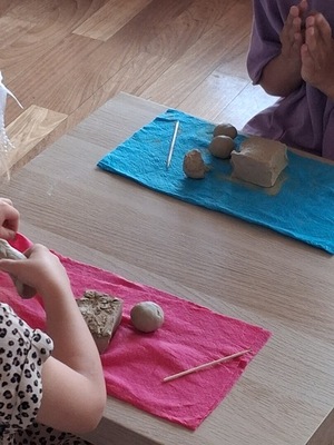 Dwoje dzieci siedzi przy stole po przeciwnych jego stronach i lepi z gliny na kolorowych podkładkch (nieieskiej i różowej). W dłoniach trzymają kawałki jasnej masy, obok leżą inne fragmenty gliny i patyczki.