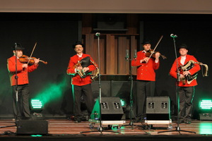 Na scenie stoją czterej mężczyźni. Ubrani w czerwone marynarki, czarne spodnie i kapelusze na głowach. W rękach trzymają instrumenty skrzypce oraz dudy. Przed nimi stoją cztery mikrofony oraz głośniki.