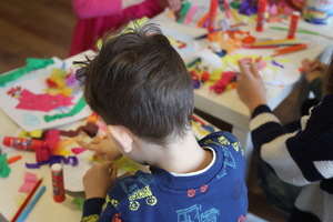 Zdjęcie z zajęć plastycznych. Na stolikach widać kolorowe kredki, kleje, krepę i inne materiały plastyczne. Na pierwszym planie chłopiec, który tworzy swoją pracę. Siedzi do nas tyłem.