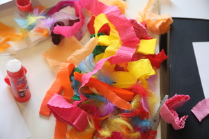 Na stole leżą kolorowe materiały plastyczne: pocięta bibuła w jaskrawych barwach, miękkie piórka oraz klej w sztyfcie, tworząc twórczy, barwny bałagan.