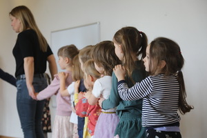 Grupa dzieci stoi w szeregu jedno za drugim. Ręce trzymają na ramionach dziecka przed sobą. Na przedzie stoi kobieta, która prowadzi dzieci. Wszyscy tworzą linię jak w zabawie lub ćwiczeniu.