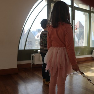 Dwoje małych dzieci stoi w jasnym pomieszczeniu przy dużym, łukowatym oknie. Dziewczynka w różowej bluzce i tiulowej spódnicy oraz chłopiec w swetrze patrzą na miasto za oknem.