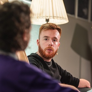 Dwie postaci siedzą obok siebie, rozmawiają. Widzimy głowy oraz część tułowia. Mężczyzna siedzi przodem. Widać twarz oraz fragment ciemnej bluzy. Druga osoba siedzi tyłem. Widać fragment sylwetki. Z mężczyzną stoi świecąca lampa.