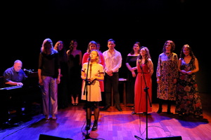 Grupa dorosłych osób stoi na scenie oświetlonej kolorowym światłem. Na pierwszym planie kobieta śpiewa do mikrofonu, za nią pozostali uczestnicy występu stoją w półkolu.