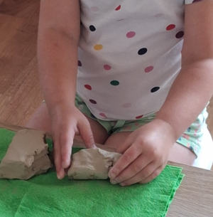 Zbliżenie na dłonie dziecka podczas warsztatów ceramicznych. Na zielonych kawałkach materiałów dziecko trzyma kawałki gliny i tworzy z niej jakiś przedmiot.