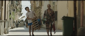 [Kolorowy kadr z filmu. Chłopiec idzie z siwą kobietą ulicą.]