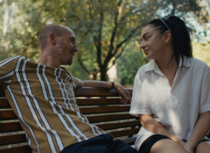 Kolorowe zdjęcie. Dwie osoby, młoda kobieta i młody mężczyzna siedzą na ławce w parku.