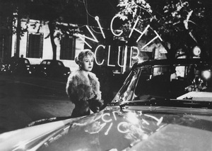 [Czarno-biały kadr z filmu. Zamyślona kobieta stoi przy samochodzie. Za nią widać neon z napisem Night Club.]