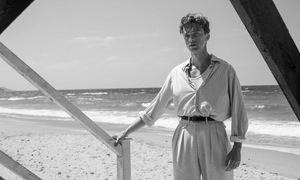 Czarno-biały kadr z filmu. Młody mężczyzna stoi na schodach. Za nim plaża i morze.