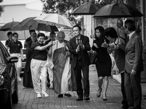 Czarno-biały kadr z filmu. Budda w szacie idzie ulicą, wokół niego tłumek ludzi, kobiet i mężczyzn w różnym wieku. Ludzie mają parasola, jedna z osób trzyma parasol nad Buddą.