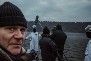 Kolorowe zdjęcie. Lewa część kadru to twarz dojrzałego mężczyzny patrzącego w kamerę. W tle nagrania filmu na pustynnym tle. Osoby w strojach kosmonautów, człowiek z mikrofonem na boomie.
