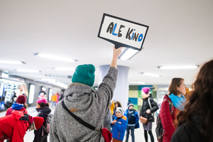 Grupa dzieci i dorosłych. Jedna osoba ma podniesioną rękę, w której trzyma tabliczkę.