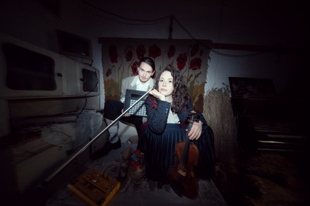 Na obrazku widać mężczyznę trzymającego muzyczną konsolę elektroniczną oraz kobietę ze skrzypcami w starej izbie.