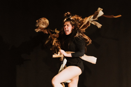 kadr z przedstawienia, skacząca kobieta, krzycząca, która trzyma w rękach kij ze szmacianą głową.