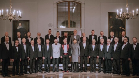 Chór Męski Copernicus - jego członkowie stojący w czarnych garniturach. Stoją w dwóch szeregach w zabytkowej sali jakiegoś pałacu. W tle duże portrety właścicieli pałacu.
