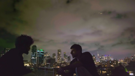 Klimatyczne, nocne ujęcie z filmu. Dwie ciemne sylwetki na pierwszym planie na dachu, zwrócone do siebie. Tło to błyszcząca panorama metropolii pod dramatycznym, zachmurzonym niebem.