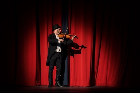 Mężczyzna w kapeluszu i długim płaszczu gra na skrzypcach, stojąc na scenie przed zasłoniętą czerwoną kurtyną, oświetlony reflektorem.