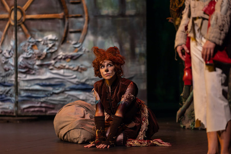 Aktorka w kostiumie kota klęczy na scenie teatralnej, w tle widoczna dekoracja z wielkim kołem i falami.