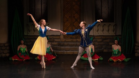 Para tancerzy baletu w kostiumach, w pozie baletowej, trzymająca się za dłonie. W tle inni uczestnicy spektaklu w bajkowych strojach, siedzący na podłodze.