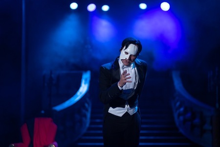 Mężczyzna w smokingu i białej masce zakrywającej pół twarzy stoi na scenie w niebieskim świetle reflektorów, wykonując dramatyczny gest dłonią.