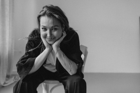 Czarno-biała fotografia uśmiechniętej, młodej kobiety. Siedzi na krześle, rękami opartymi na kolanach podtrzymuje głowę. Ma długie, rozpuszczone włosy.