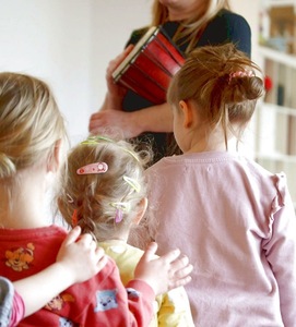 Trójka dzieci stoi plecami do aparatu i patrzy na nauczycielkę w tle, która trzyma mały bębenek. Dziewczynka w środku ma we włosach kolorowe spinki. Lewa dłoń dziecka opiera się na ramieniu sąsiadki. Zdjęcie ma miękkie, naturalne oświetlenie.