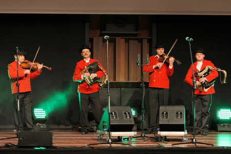 Na scenie stoją czterej mężczyźni. Ubrani w czerwone marynarki, czarne spodnie i kapelusze na głowach. W rękach trzymają instrumenty skrzypce oraz dudy. Przed nimi stoją cztery mikrofony oraz głośniki.