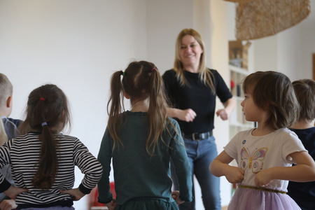 Grupa dzieci stoi w półkolu z rękami opartymi na biodrach, patrząc na uśmiechniętą instruktorkę, która pokazuje im ruchy podczas zajęć.