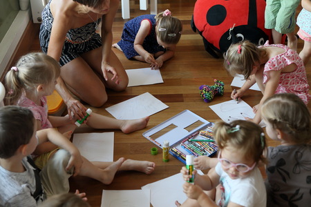 Grupa dzieci rysuje na podłodze na białych kartkach. Siedzą w kole. Podłoga jasne, drewniana. W tle osoba prowadząca oraz duża pluszowa poduszka w kształcie biedronki owada.