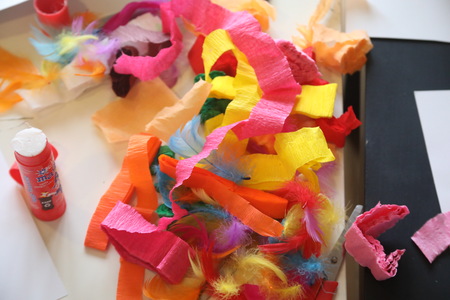 Na stole leżą kolorowe materiały plastyczne: pocięta bibuła w jaskrawych barwach, miękkie piórka oraz klej w sztyfcie, tworząc twórczy, barwny bałagan.