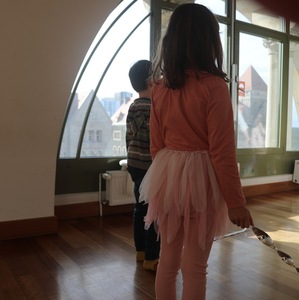 Dwoje małych dzieci stoi w jasnym pomieszczeniu przy dużym, łukowatym oknie. Dziewczynka w różowej bluzce i tiulowej spódnicy oraz chłopiec w swetrze patrzą na miasto za oknem.